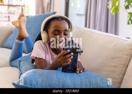 Jeune femme afro-américaine allongée sur un canapé, portant un casque crème et tenant un smartphone souriant. Casque, appareil, canapé, fenêtre, lumière du soleil, usine, Banque D'Images