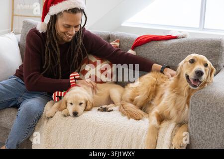 Souriant homme mi-adulte portant chapeau de Père Noël caressant deux chiens dorés sur le canapé gris, jouet en peluche de bonbons. Animaux de compagnie, confortable, festif, lumière du jour, chaleur, compagnonnage, h Banque D'Images