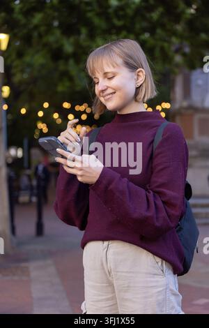Femme debout dans la rue au crépuscule portant un pantalon marron de bronzage pull tenant le sac à dos de smartphone. Urbain, crépuscule, place, arbres, lumières, bokeh, téléphone Banque D'Images