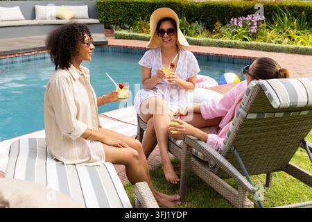 Différentes amies femmes discutant tout en tenant des cocktails garnis au bord de la piscine dans le jardin, portant des lunettes de soleil. Chapeau de soleil, lunettes de soleil, extérieur Banque D'Images