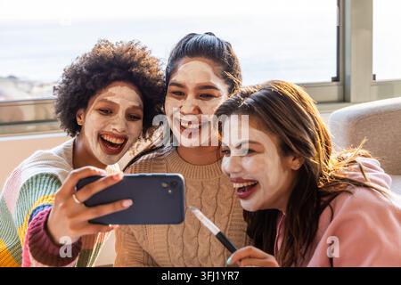 Diverses amies féminines assis sur le canapé près de la fenêtre portant des masques de visage posant avec téléphone et brosse. Selfie, soins de la peau, copines, confortable, canapé, dayligh Banque D'Images