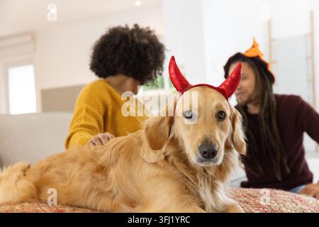 Couple caressant Golden retriever portant des cornes rouges sur canapé femme moutarde pull homme orange chapeau. Chien, bandeau, canapé, maison, salon, famille, natura Banque D'Images