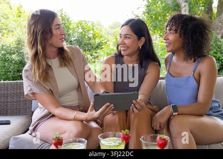 Trois amies diverses assises côte à côte sur un canapé de terrasse, regardant une tablette et sirotant des boissons vertes. Extérieur, arrière-cour, femmes, c Banque D'Images