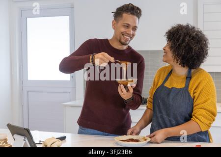 Couple, femme dans le tablier et homme dans le pull en tricot, préparant la tarte à l'îlot de cuisine avec bol. Duo, homecooking, pâtisserie, collaboration, cosy, brick Banque D'Images