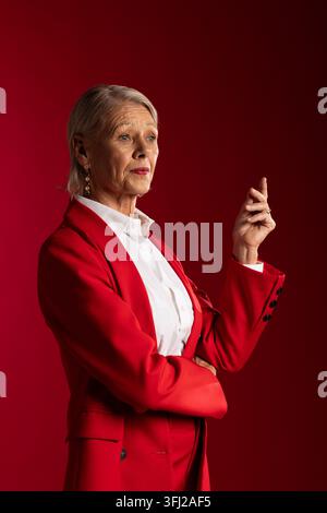 Femme âgée posant en studio, portant un blazer rouge et un pantalon, une chemise blanche, des boucles d'oreilles en forme de goutte et une bague Banque D'Images