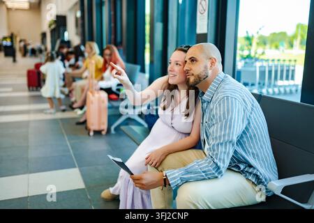 Jeune couple assis dans un salon d'aéroport, pointant et regardant quelque chose, tenant des cartes d'embarquement Banque D'Images