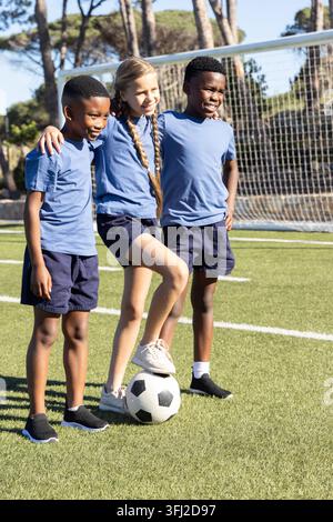 Divers enfants reliant les bras sur le terrain de football, pied sur le ballon de football par but portant un kit bleu ciel. Coéquipiers, jeunesse, balle, gazon, filet, soleil, ombres Banque D'Images