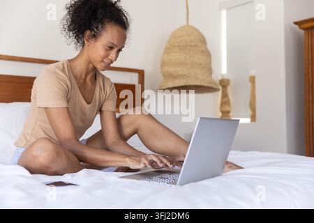 Femme afro-américaine et asiatique assise sur un lit en train de taper sur un ordinateur portable argenté à côté d'un smartphone à la maison. Minimaliste, confortable, moderne, ser Banque D'Images