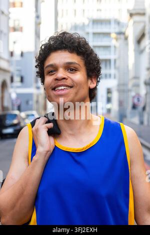 Afro-Américain début de la vingtaine debout dans une rue urbaine tenant une serviette noire, en maillot bleu et jaune. Athlète, paysage urbain, lumière du jour Banque D'Images