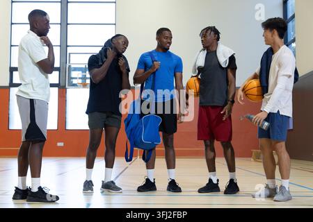 Divers coéquipiers masculins dans l'équipement de gymnastique bavardant sur le terrain de gymnastique, tenant des ballons de basket, sac de sport, serviettes. Athlètes, travail d'équipe, décontracté, installation, dynamique, focu Banque D'Images