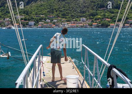 4 juillet 2026, Kotor, Monténégro, Un garçon vêtu de vêtements d'été décontractés se tient sur une pente descendant vers l'eau dans un port, devant une eau bleu clair Banque D'Images