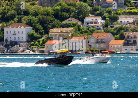 4 juillet 2026, Kotor, Monténégro, Deux motos nautiques filent sur une eau bleue et calme, laissant des sillage blancs éclatants alors que le village de bord de mer en colline avec des maisons en terrasses Banque D'Images