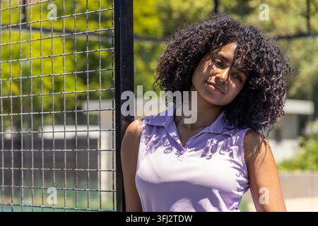 Femme afro-américaine appuyée contre un poteau métallique près d'une clôture, la lumière du soleil projetant des ombres sur un polo lilas. Extérieur, parc, ensoleillé, décontracté Banque D'Images