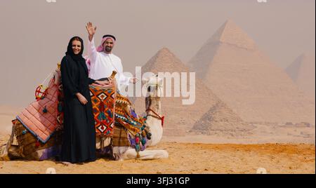 Un couple arabe en tenue traditionnelle pose avec un chameau décoré aux pyramides de Gizeh, l'homme agitant joyeusement la main. Autorisations de modèle sur fichier. Idéal pour les voyages au Moyen-Orient et en Afrique du Nord Banque D'Images