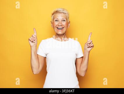 Portrait d'une femme senior aux cheveux courts montrant un geste de pouce levé et souriant, isolée sur un fond jaune Banque D'Images