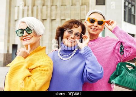 Trois amies âgées joyeuses et heureuses portant des pulls colorés et des lunettes de soleil marchent ensemble près d'un bâtiment moderne. Banque D'Images