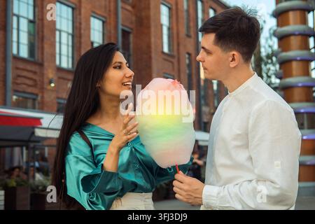 Adorable couple amoureux se promène en ville et mange de la barbe à papa. Jeune femme asiatique aux cheveux longs et jeune homme européen. Banque D'Images
