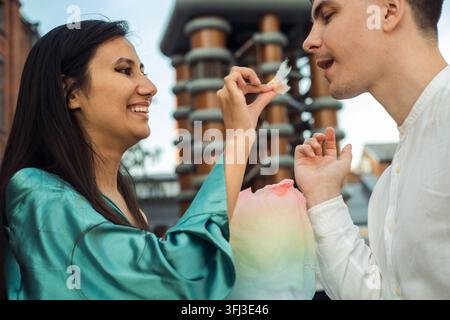 Adorable couple amoureux se promène en ville et mange de la barbe à papa. Jeune femme asiatique aux cheveux longs et jeune homme européen. Banque D'Images