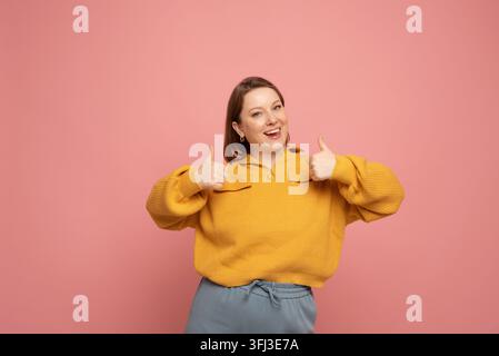 Portrait d'une femme ronde en pull jaune montrant un geste de pouce levé, isolé sur fond rose. Banque D'Images