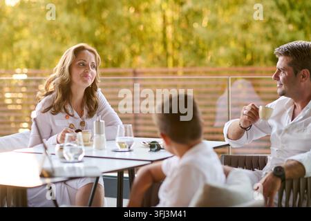 Un moment familial détendu sur une terrasse ensoleillée, alors que les parents et l'enfant profitent d'un café et d'une conversation avec une vue mer chaleureuse en toile de fond, capturant la complicité Banque D'Images
