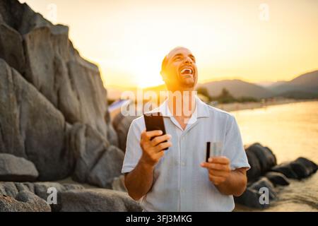 Homme riant en utilisant un téléphone portable et une carte de crédit pour acheter en ligne Banque D'Images