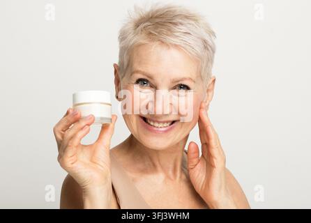 Une charmante femme âgée aux cheveux courts sourit et tient un pot de crème dans ses mains. Routine de beauté. Banque D'Images
