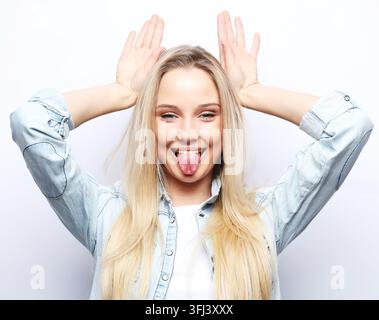 Jeune femme blonde en tenue décontractée faisant un geste amusant avec le doigt au-dessus de la tête comme des cornes de taureau Banque D'Images