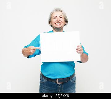 Une femme âgée heureuse et charmante tient une feuille de papier vierge dans ses mains et pointe du doigt dessus. Isolée sur fond blanc. Banque D'Images