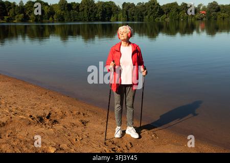 Une femme pensionnée heureuse et âgée est engagée dans la marche nordique avec des bâtons et écoute de la musique avec des écouteurs. Banque D'Images