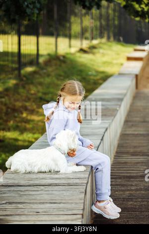 Une fille de sept ans avec des couettes est assise sur un banc du parc et joue avec un chien blanc Banque D'Images