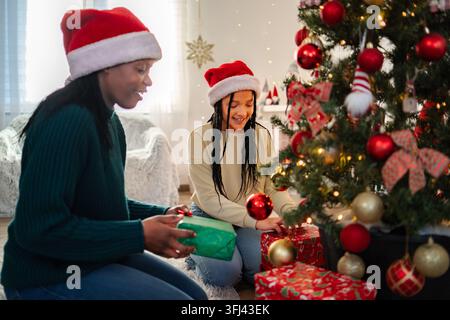 Mère et fille portant des chapeaux du Père Noël profitant de la tradition des fêtes, décorant le sapin de Noël avec des cadeaux Banque D'Images