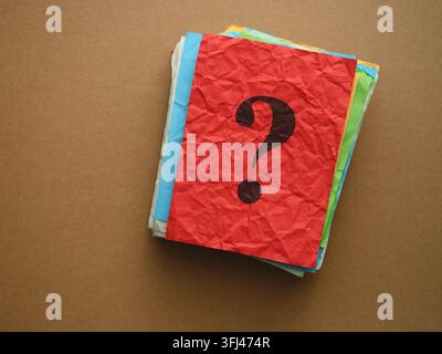 Vue de dessus d'un point d'interrogation sombre imprimé sur un morceau de papier rouge froissé posé sur une pile de papiers colorés contre un fond marron. Banque D'Images