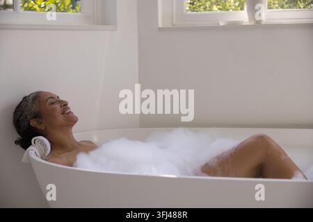 Femme allongée dans une baignoire indépendante dans la salle de bain à la maison, entourée de bulles blanches denses. Minimaliste, lumière du jour, calme, spa, verdure, mousse, neutre Banque D'Images
