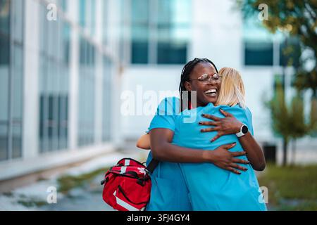 Deux professionnelles de santé féminines diverses s'embrassant joyeusement devant un hôpital Banque D'Images