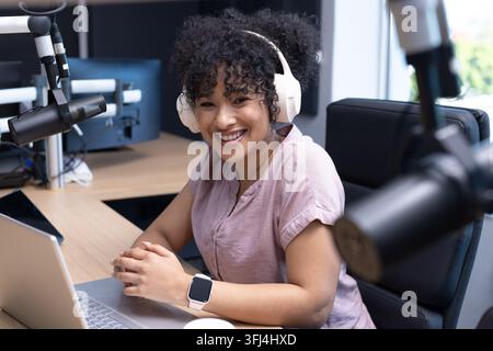 Femme afro-américaine assise à un bureau de studio portant des écouteurs blancs, utilisant un ordinateur portable avec des microphones à perche Banque D'Images
