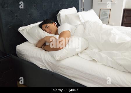 Homme asiatique dormant sur un lit en T-shirt blanc, serrant un oreiller près de la tête de lit capitonnée. Chambre, repos, sérénité, luxe, literie, natu Banque D'Images
