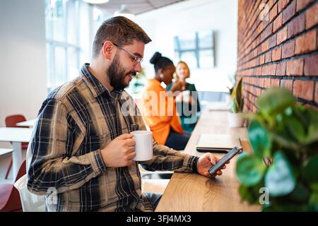 Homme utilisant un smartphone et buvant du café pendant la pause au bureau Banque D'Images