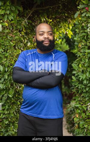 Homme debout au centre d'une arche de haie, portant une chemise de sport bleue et des manches de compression noires. Verdure, chemin, jardin, parc, barbu, portrait, ou Banque D'Images