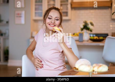 Femme enceinte heureuse goûtant un dessert de boulangerie dans une cuisine Banque D'Images