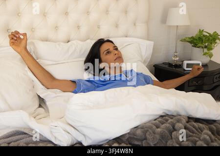 Indienne allongée sur le lit dans la chambre, portant un haut de pyjama en satin bleu, atteignant le réveil numérique. Tufté, serein, confortable, texture Banque D'Images