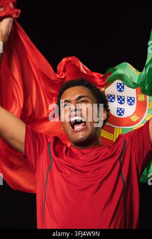 Homme afro-américain portant une chemise rouge, agitant le drapeau national avec emblème et criant en studio. Bannière, cramoisi, émeraude, gros plan, portrait, fond sombre Banque D'Images