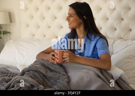 Indienne assise dans le lit tenant une tasse en terre cuite, portant un haut de pyjama en satin bleu. Chambre, confortable, sereine, matin, relaxation Banque D'Images