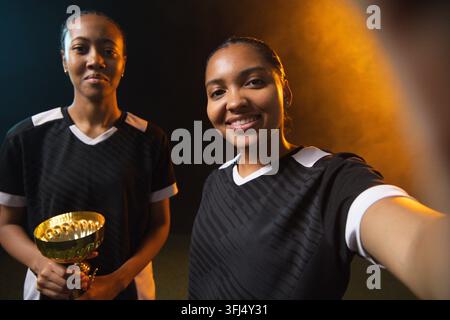 Athlètes afro-américaines en maillots prenant un selfie en studio, tenant un trophée d'or et un téléphone. Portrait, coéquipières, célébration Banque D'Images