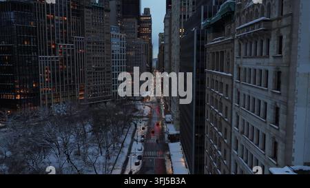 Vol lent en drone au-dessus d'une rue enneigée à New York à la tombée de la nuit, avec la circulation se déplaçant entre les immeubles de grande hauteur, les appartements et les tours de bureaux créant Banque D'Images