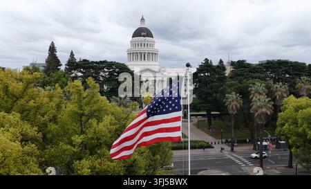Vue aérienne par drone du drapeau des États-Unis flottant devant le Capitole de l'État de Californie, entouré d'arbres et d'architecture civile à Sacramento, Californie Banque D'Images