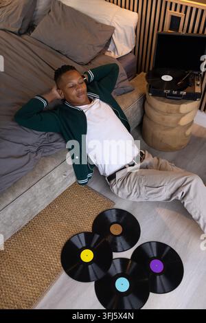 Homme africain dans la vingtaine allongé sur le sol de la chambre à côté d'un lit bas, platine vinyle en lecture Banque D'Images