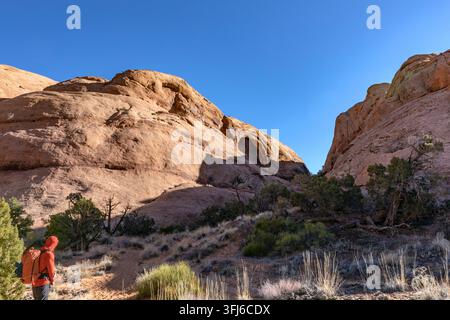 Randonneur en veste orange avec un sac à dos marche vers une arche de grès rouge dans Bow and Arrow Canyon près de Moab, Utah, États-Unis sous un ciel bleu clair Banque D'Images