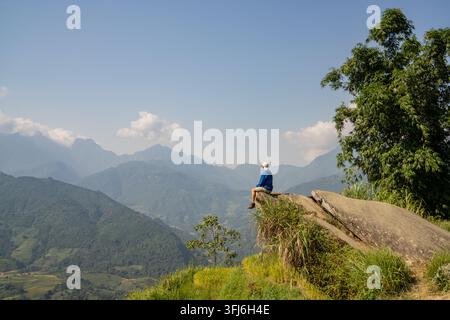 Homme assis sur le bord d'une roche avec les jambes pendantes au-dessus d'une vallée montagneuse, Y Ty, Lao Cai, Vietnam Banque D'Images