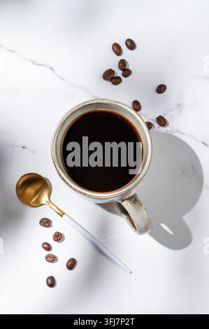 Café espresso fraîchement préparé dans une tasse sur un fond de marbre blanc avec des grains de café éparpillés, une cuillère et la lumière du matin. Un petit-déjeuner délicieux co Banque D'Images