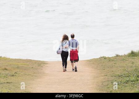 Vue arrière d'un couple les bras autour de l'autre marchant le long d'un sentier côtier au sommet d'une falaise en direction de la mer. Banque D'Images